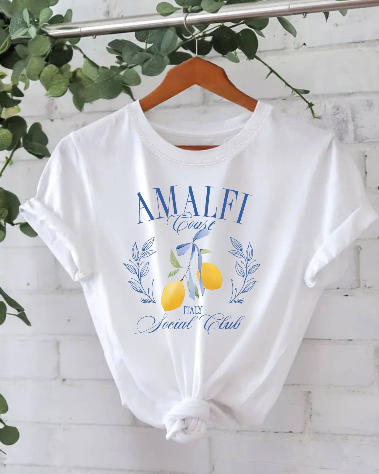 The Stylish Lemon Graphic Print Crew Neck T-Shirt by Rosa Clothing features an Amalfi Coast lemon design and "AMALFI Coast ITALY Social Club" text, displayed on a hanger against a white brick wall with green foliage.