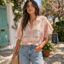 A woman in a Pine Apparel Chiffon Embroidered Button Front Blouse and jeans stands outdoors near green doors and flowers, with a "Fresh flowers & coffee" sign visible in the background.