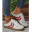 A person wears FASHION GO’s Mesh Low Back Slip On Sneakers—cream slip-ons accented with red chevron stripes, white laces, and subtle mesh panels for breathability—matched with frayed hem blue jeans and a gold anklet, standing on a gray surface near wooden planks and green plants.