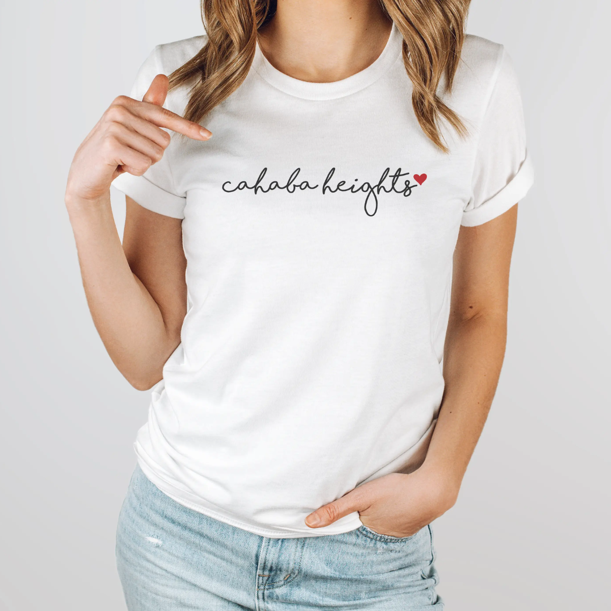 A woman wears the Faire Custom Local Love Script Graphic Tee, a modern fit cotton-poly blend with "cahaba heights" and a small red heart printed on it, as she points toward the text.