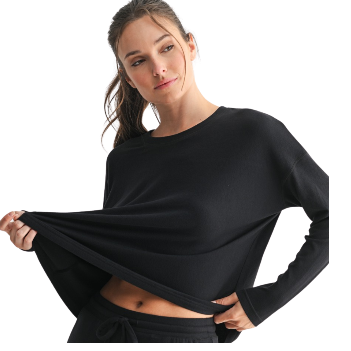 A woman wearing the Mono B Brushed Hacci Long Sleeve Top holds out the bottom of her black shirt, showing part of her midriff.