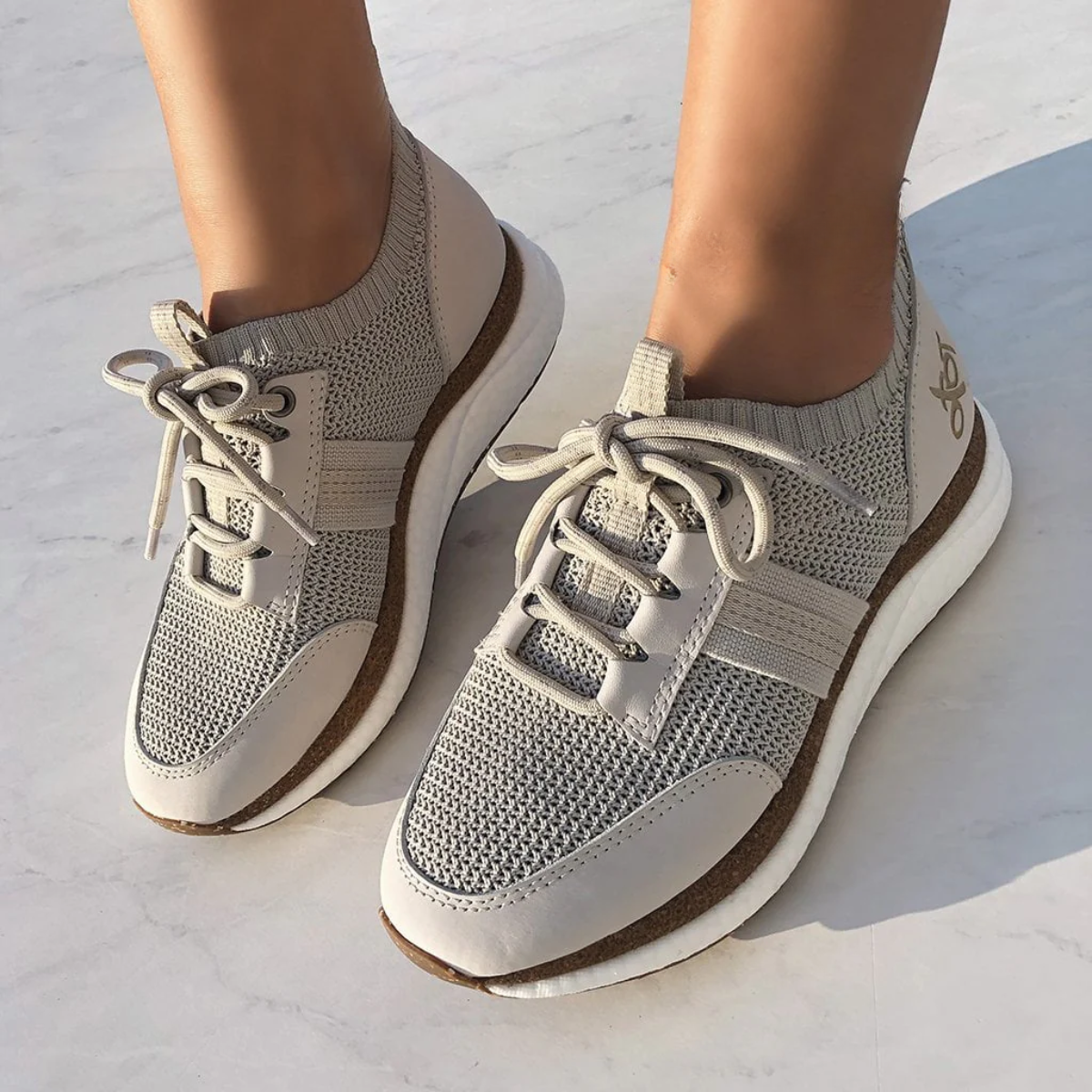 A person wears Speed Sneakers in Mist by OTBT from Consolidated Shoe Co—light gray women's sneakers with white laces and a recycled rubber sole—standing on a light-colored surface.