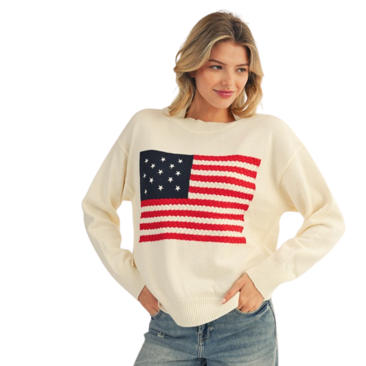 A woman in a cozy, cream-colored FASHION GO American Flag Sweater and blue jeans stands with her hands in her pockets, radiating all-American charm.
