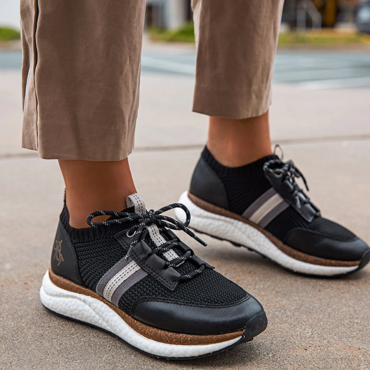 A person in beige pants stands outdoors on concrete, wearing the Speed Sneakers in Black by OTBT from Consolidated Shoe Co, featuring gray stripes and white recycled rubber soles for added bounce back technology.