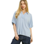 A woman wears the Umgee Linen Frayed Edge Short Sleeve Top in light blue with a split neck, paired with dark jeans, standing against a white background.