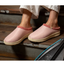 A person wearing COBIAN Nami Flip Slips in light pink, featuring wavy stitching and an EVA cupsole, stands on wooden steps. Only their lower legs and feet are visible, highlighting this closed-toe comfort footwear.