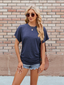 A woman in a Suhkasana Relaxed Fit Short Sleeve Rayon Blend Tee for women, navy blue, paired with denim shorts, sunglasses, and a brown shoulder bag stands before a brick wall.