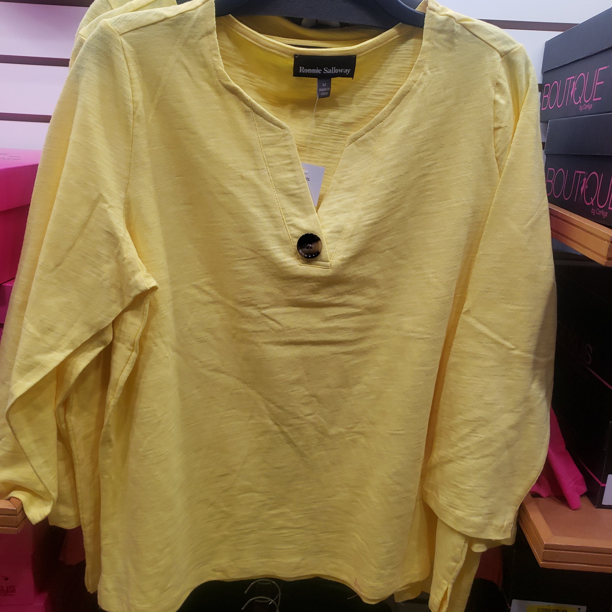 A 3/4 Sleeve Button Accent Cotton Top in Yellow from RONNIE SALLOWAY & CO INC is displayed on a hanger in a clothing store.