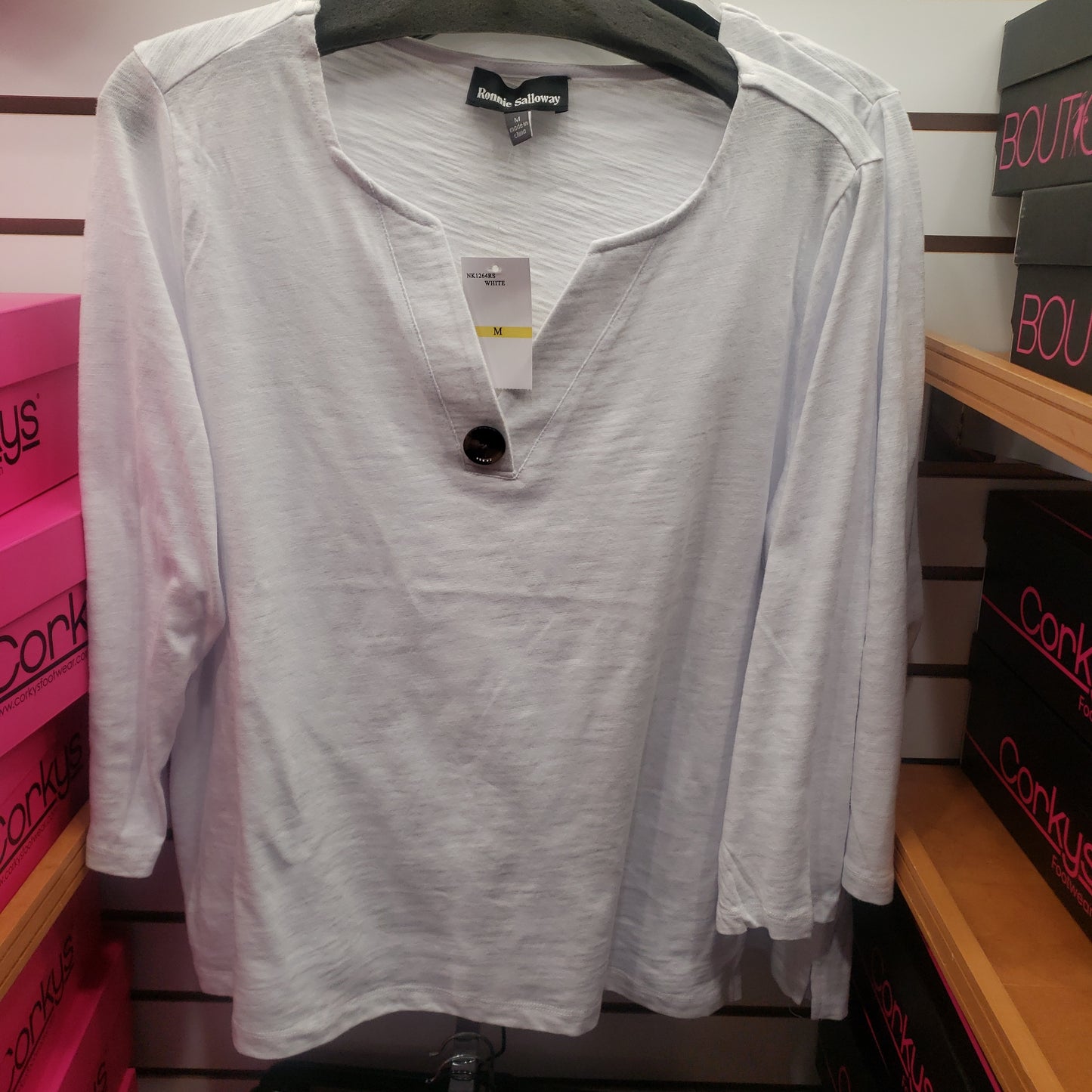 A RONNIE SALLOWAY & CO INC 3/4 Cotton Top with Button Accent in White hangs on a rack. Pink and black boxes are visible on the shelves in the background.