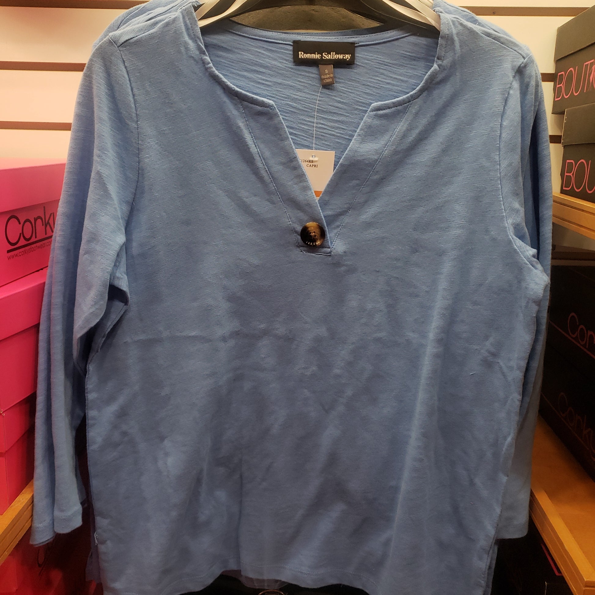 A 3/4 Sleeve Wood Button Accent Cotton Top in Sky Blue by RONNIE SALLOWAY & CO INC hangs on a store rack. Multiple shoeboxes are visible in the background.