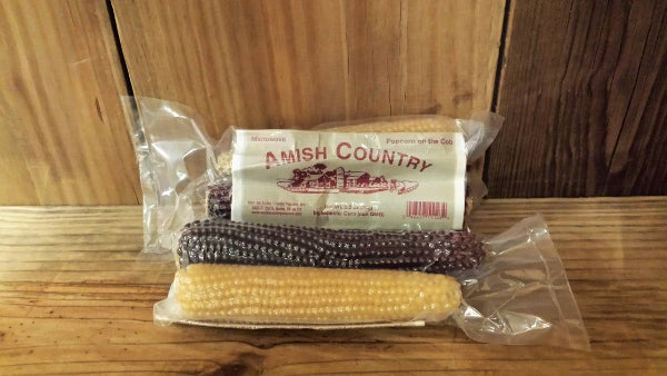 Two vacuum-sealed Twin Microwave Popcorn Cobs from Amish Country Popcorn, with colorful kernels, are displayed on a wooden surface against a wooden background.