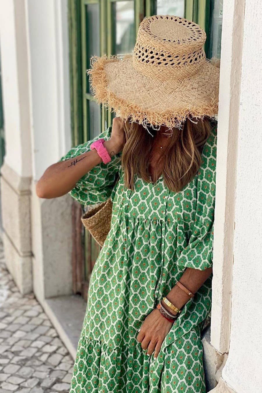 A person in a Rosa Clothing Bohemian Floral Balloon Sleeves loose flare swing dress and a large straw hat leans against a white wall, partially covering their face with the hat.