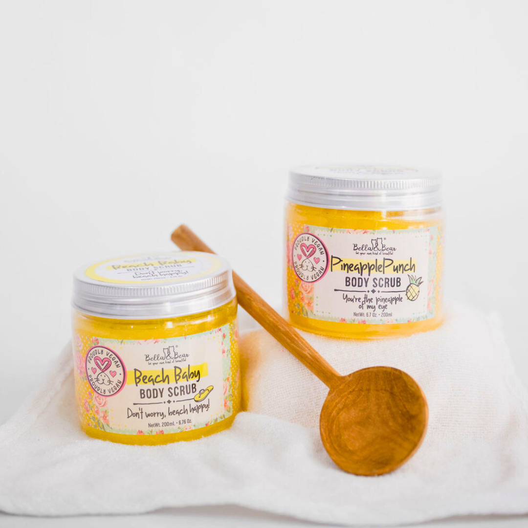 Two jars of Bella & Bear 6.7oz Beach Baby Body Scrub with a wooden spoon rest on a white towel, offering a beach glow and tropical escape vibe against a simple background.