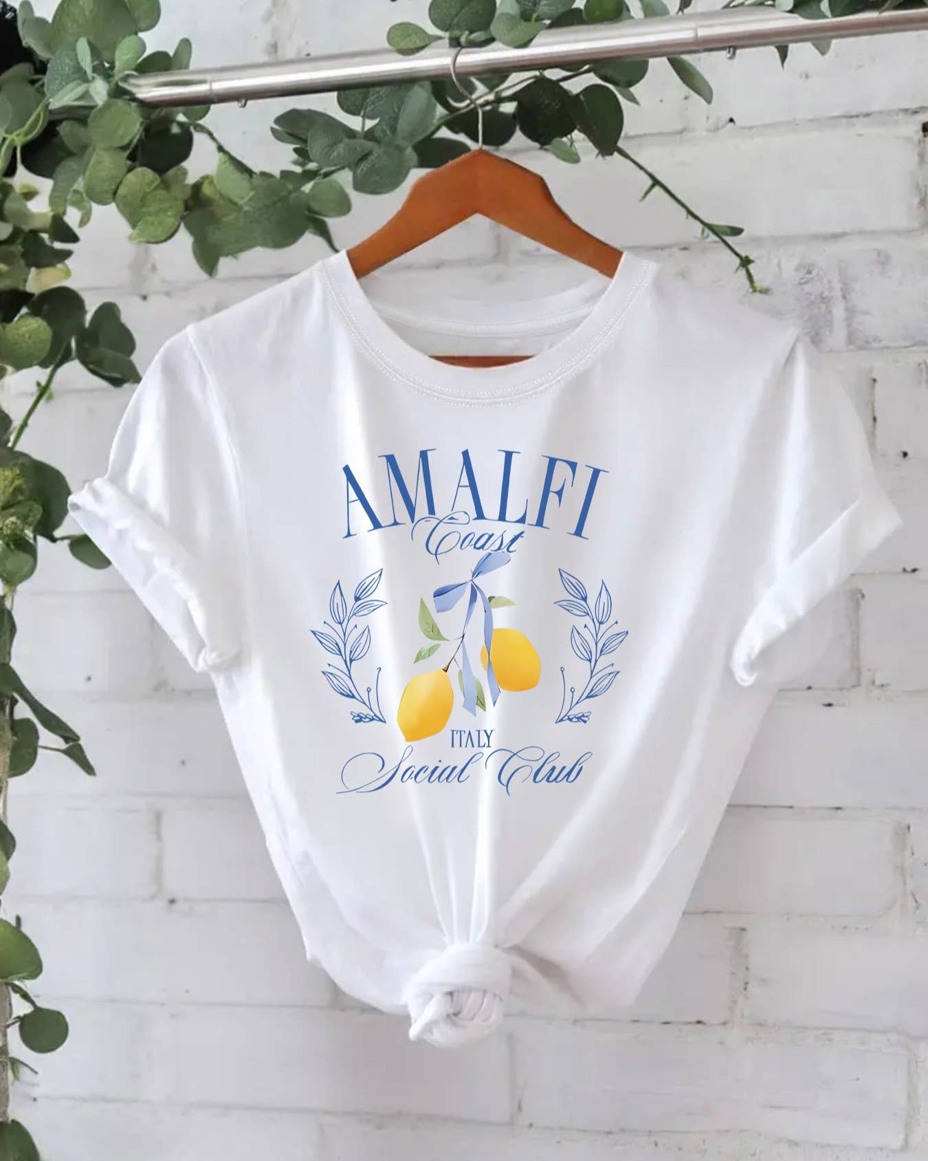 The Rosa Clothing Stylish Lemon Graphic Print Crew Neck T-Shirt features "Amalfi Coast Italy Social Club" text and a lemon with laurel illustrations on the front, shown in white on a hanger.