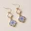 Lilla Haven’s Blue Enamel Clover Drop Earrings feature a graceful quatrefoil shape adorned with a blue floral motif on a white enamel background, finished with elegant gold trim and shown resting on a neutral surface.