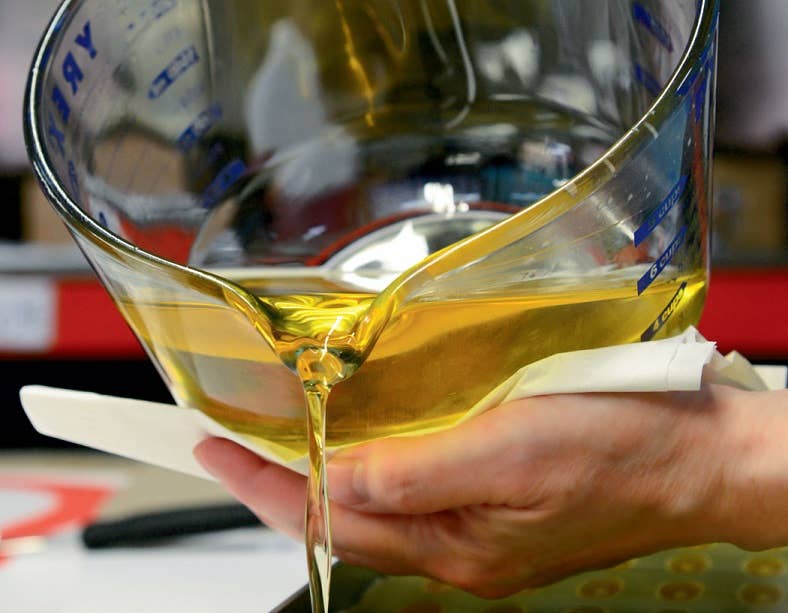 With a measuring cup, someone pours the vibrant yellow liquid of PUREfactory Naturals' The Works Travel Size Skin Care Favorites - Orange Honey with Orange Essential Oil, possibly crafting it for skin repair.