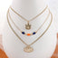 Discover the Auburn Gold & Tube Bead Trio Necklace Set by Seasons Jewelry: a stunning three-layered necklace displayed on a stand. The top layer features an "A" pendant, the middle showcases orange and blue tube beads inspired by Auburn University designs, and the bottom displays an oval medallion with an intricate design—a perfect War Eagle jewelry statement piece.