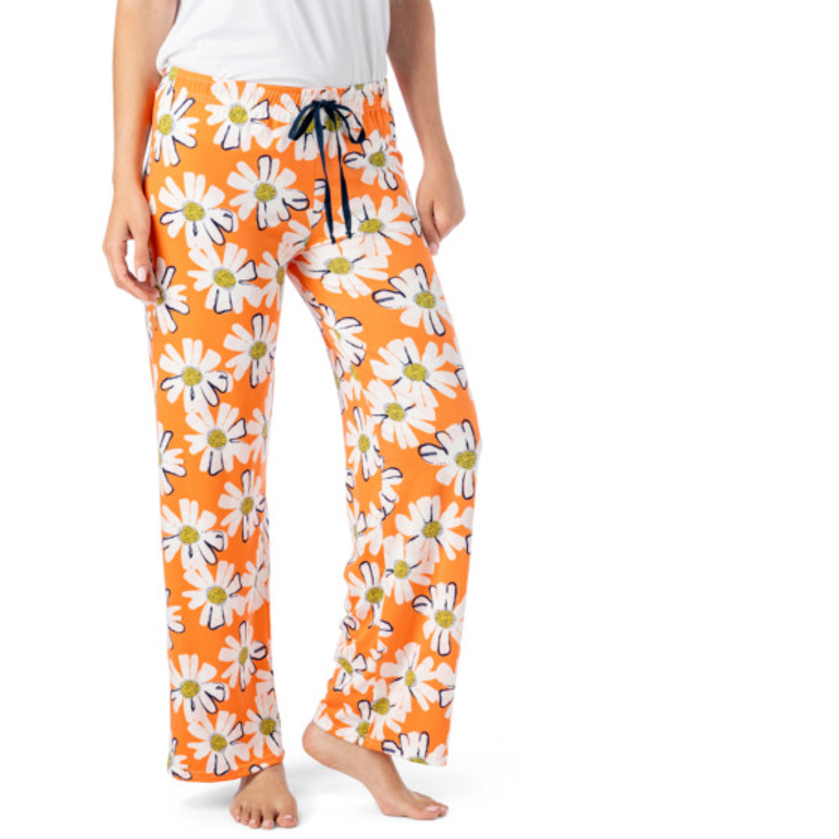 A person, barefoot and wearing the Hello Mello Lounge Pants Spring Edition by DM MERCHANDISING INC, is dressed in orange pajama pants featuring a white daisy pattern and a relaxed wide-leg fit. Paired with a simple white shirt, these stretchy pants offer enhanced comfort and ease.