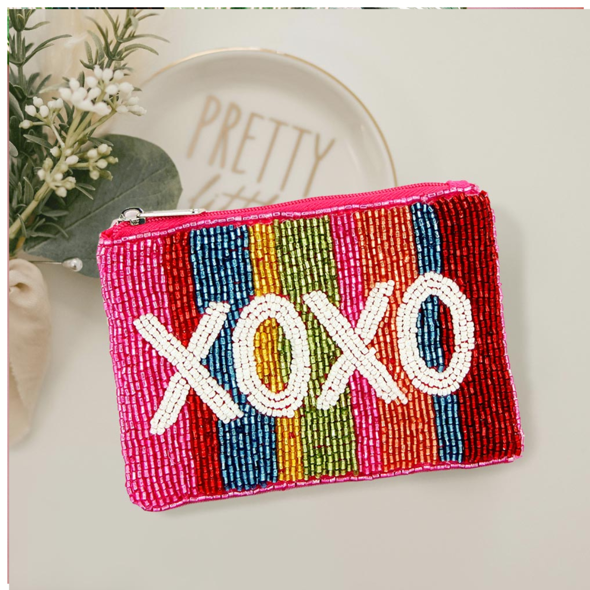The Wona Trading Seed Bead Accented Mini Pouch Bag Assorted features vertical multicolored stripes and "XOXO" in white beads—an ideal gift for fashionistas, shown on a white surface beside a "PRETTY" plate and greenery.