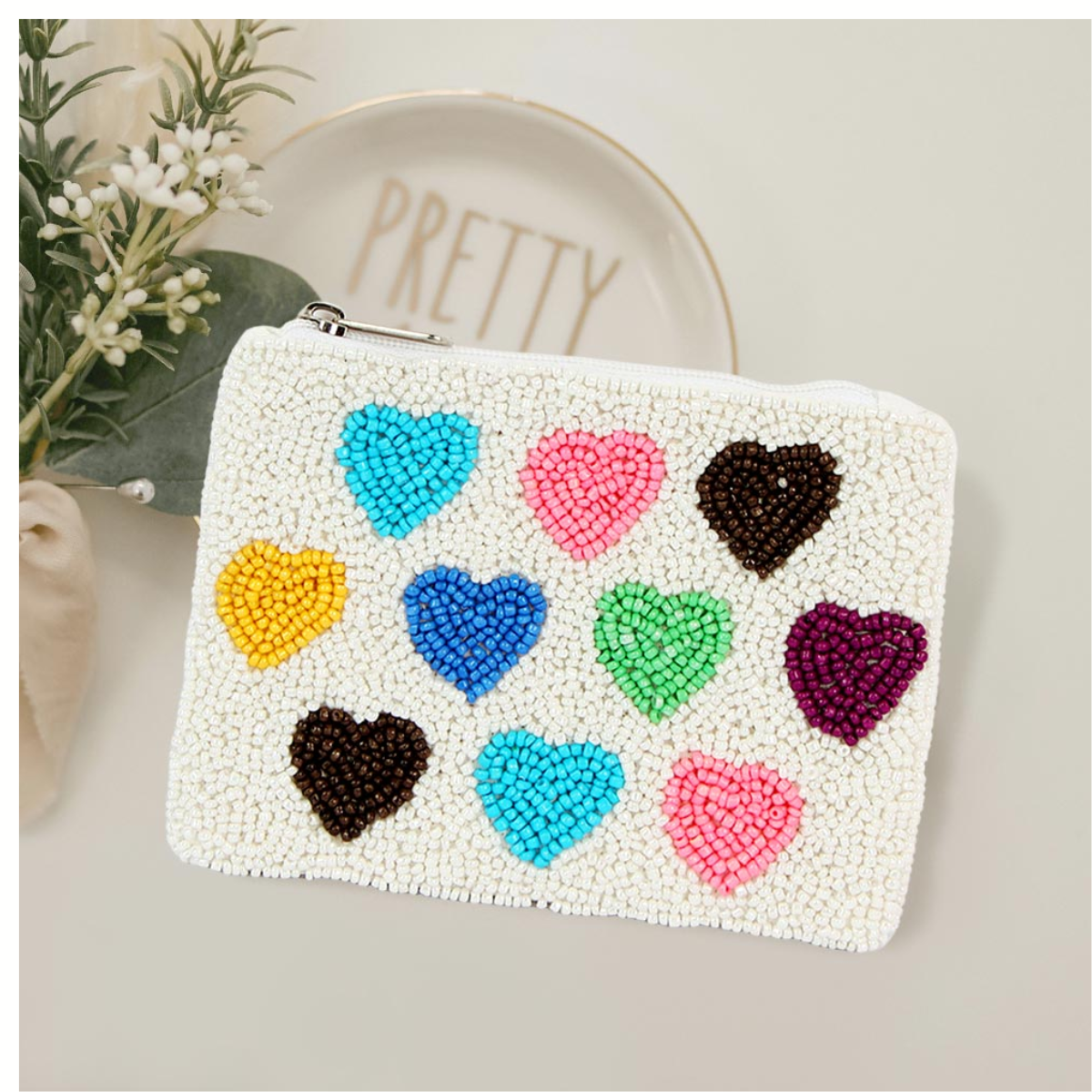 The Seed Bead Accented Mini Pouch Bag Assorted by Wona Trading features white seed beads with nine colorful hearts in rows—styled beside a decorative plate and greenery, it’s a perfect gift for fashionistas.