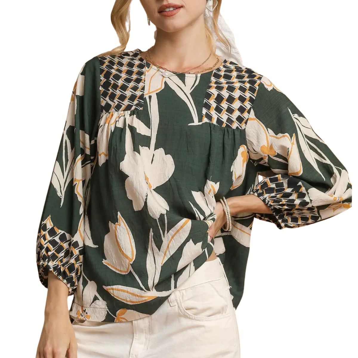 A woman wears the Umgee Mix Floral Print Blouse with Contrast Yoke, featuring long sleeves, dark green floral patterns, and white and yellow geometric details, styled with light-colored pants.
