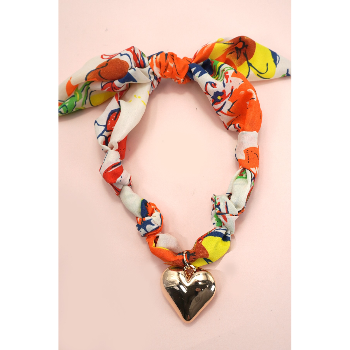 The FASHION GO Handmade Scarf Necklace in assorted colors and patterns features a bold metallic gold heart charm at the center, styled like a trendy statement piece, and is displayed on a light pink background.