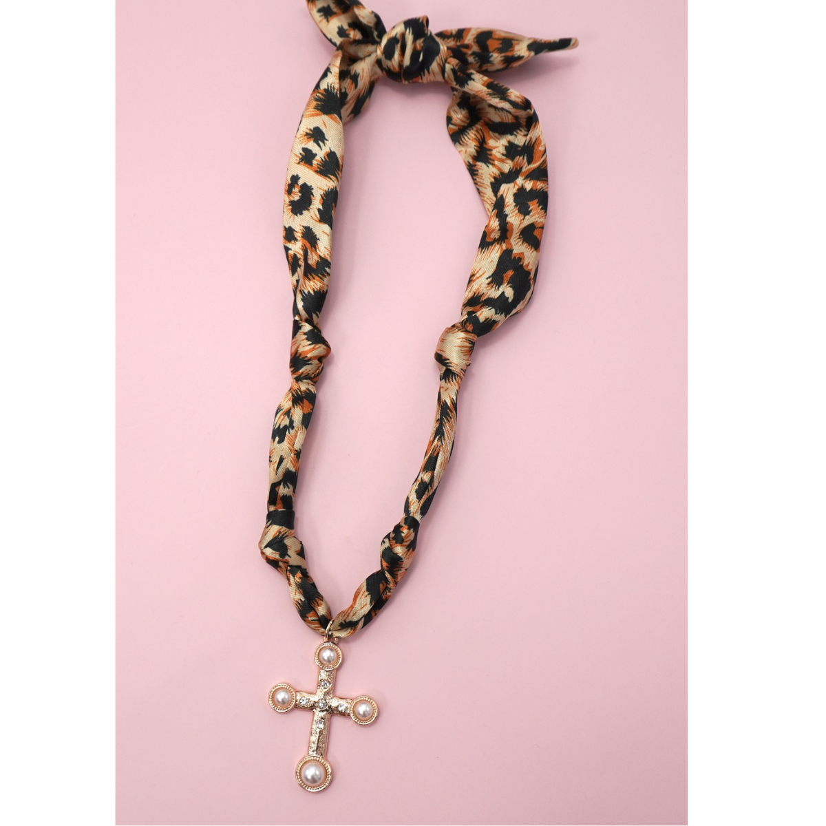 The FASHION GO Handmade Scarf Necklace in assorted colors and patterns features on-trend prints with unique charms, including a gold and pearl cross, shown against a pink background.