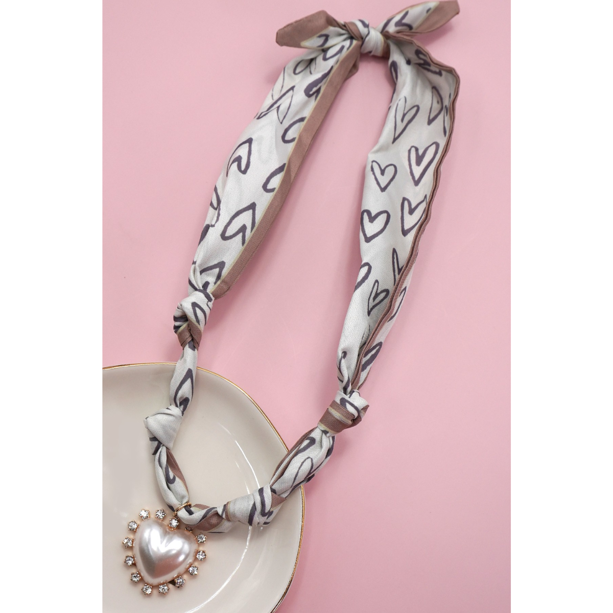 The FASHION GO Handmade Scarf Necklace—available in assorted colors and patterns—is styled with a rhinestone heart-shaped pendant and displayed on a white dish against a pink background.