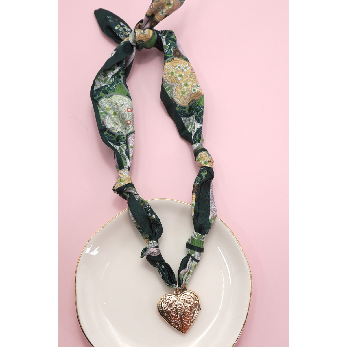 The FASHION GO Handmade Scarf Necklace, shown in a green patterned fabric with assorted charms including a gold heart-shaped locket, is displayed on a white plate against a pink background.