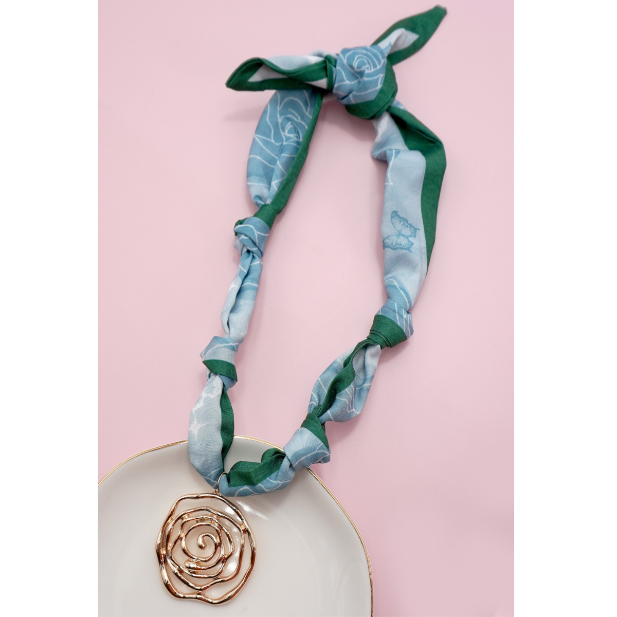 The FASHION GO Handmade Scarf Necklace in assorted colors and patterns, shown on a pink surface with a gold rose-shaped pendant, brings vibrant charm to any outfit.