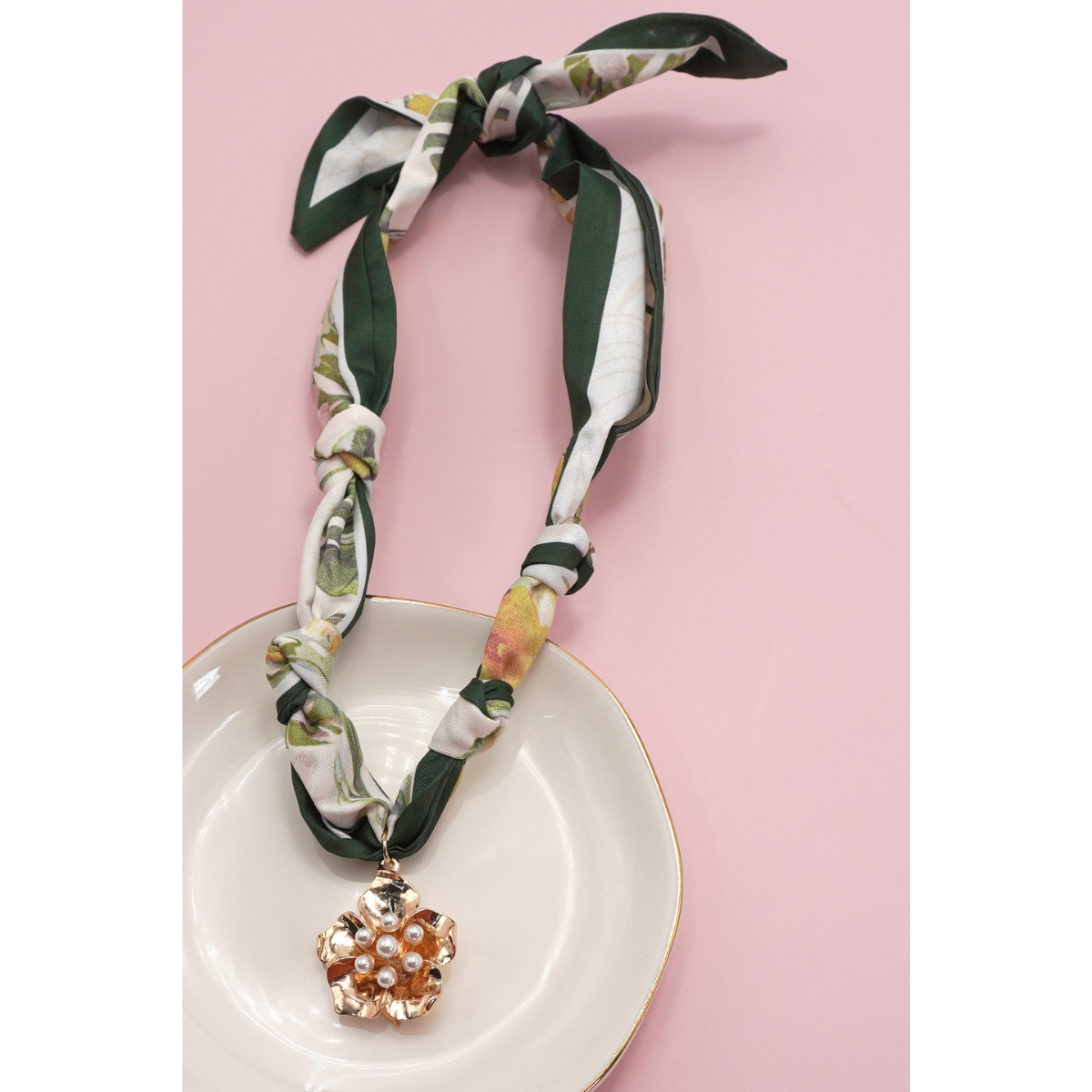 The FASHION GO Handmade Scarf Necklace in assorted colors and patterns, adorned with charms and a gold pendant with pearl accents, is showcased on a white dish against a pink background.