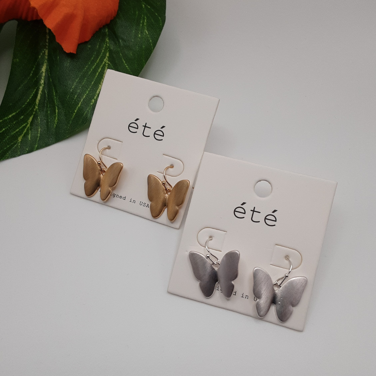 Two pairs of "Drop Butterfly Earring Matte Metal" by SPECIAL EFFECTS are displayed on white cards labeled âÊtÊ.â One pair is gold-toned, and the other is silver-toned. A green leaf and an orange flower can be partially seen in the background.