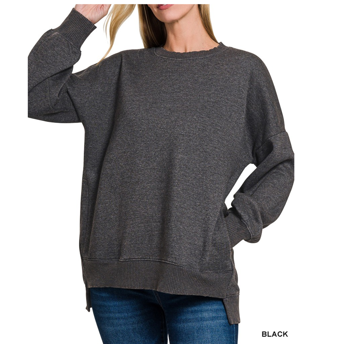 A woman wears a loose-fitting Zenana Acid Wash Fleece Hi-Low Hem sweatshirt in dark gray and blue jeans, one hand in her pocket, the other touching her hair. The text "BLACK" is visible in the lower right corner.