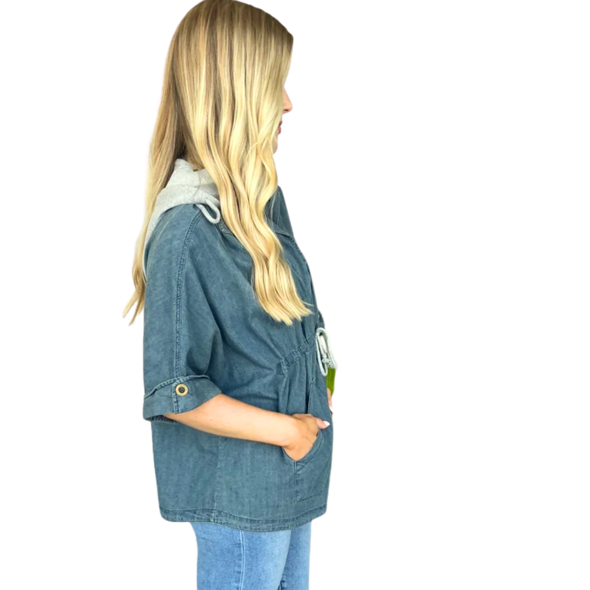 A woman with long blonde hair stands in profile, wearing the Carole Christian Denim Shirt Jacket with Detachable Hood and rolled sleeves, paired with light blue jeans.
