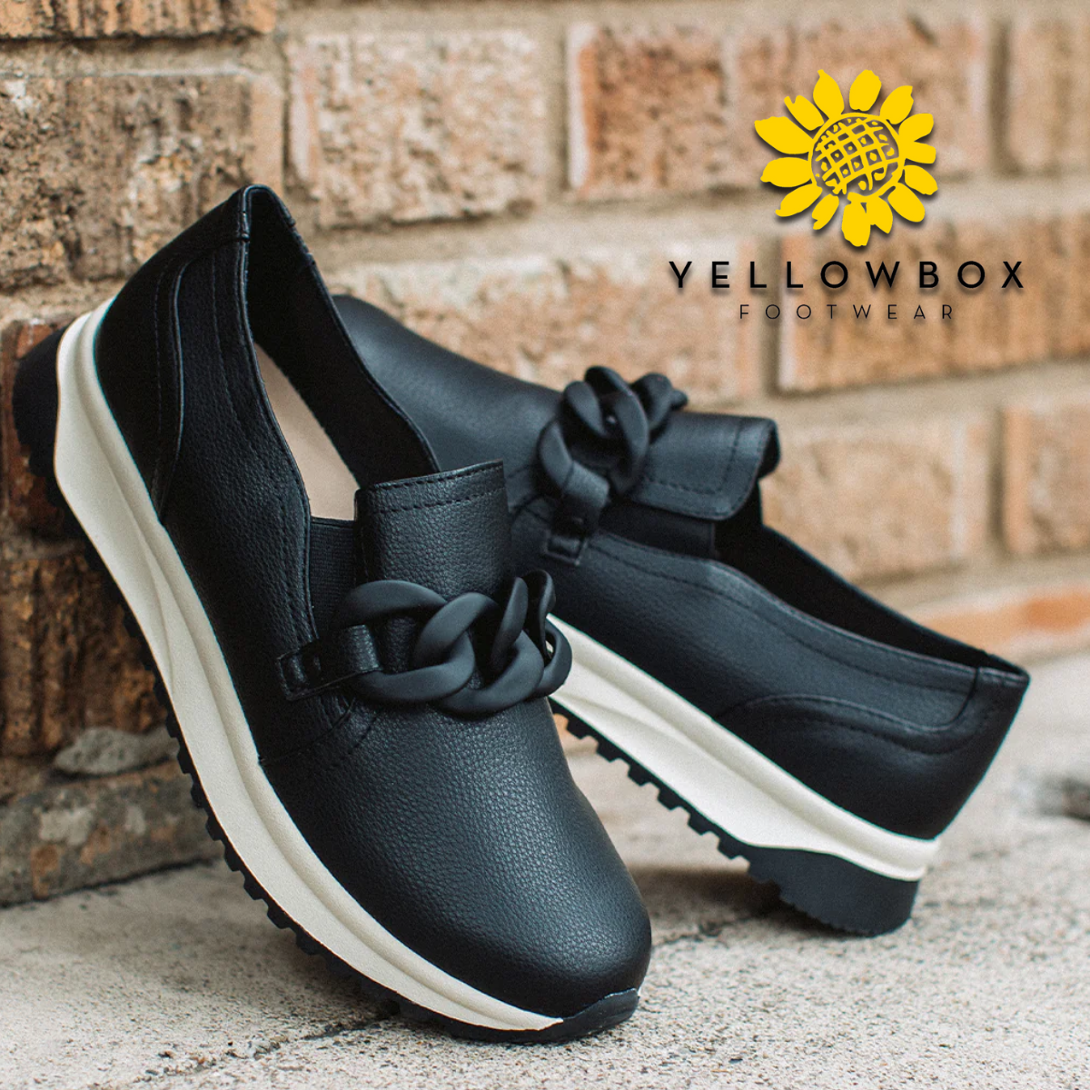 The Black Rory Loafers by Yellow Box, featuring chic slip-on design and chunky chain hardware, are set against a brick wall. The YELLOW BOX - CIT logo, adorned with a sunflower, is visible in the top right corner.