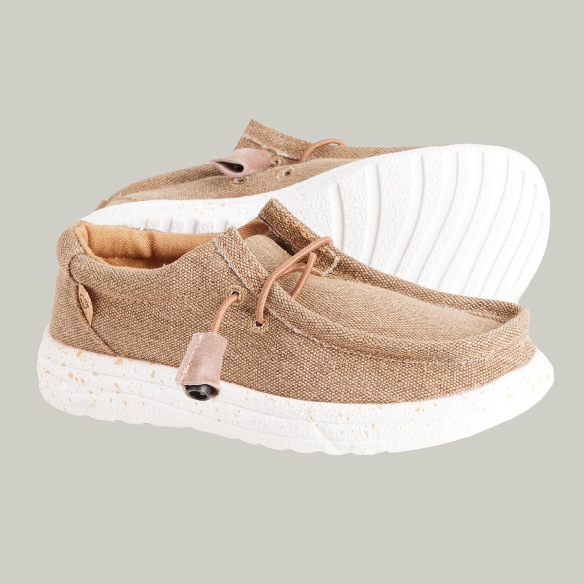 The Paulie Kids Shoe in Khaki by LAMO boasts durable beige canvas with textured fabric, white speckled soles, and light brown toggle fasteners. Ideal on gray backgrounds, they offer comfort with cushioned insoles.
