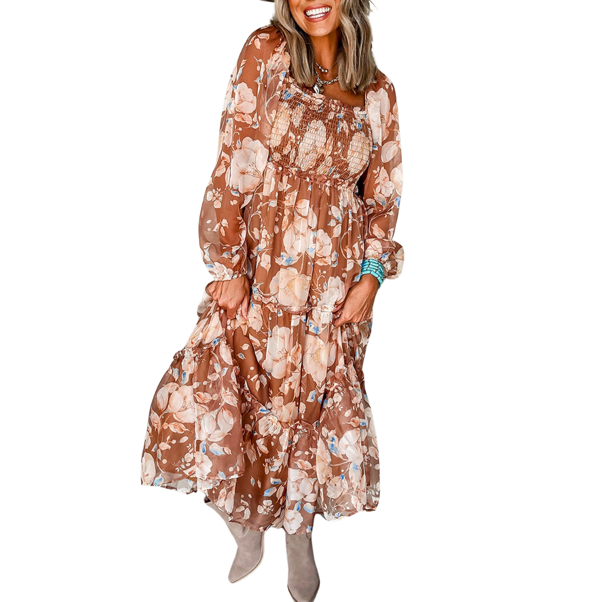 A person is smiling while wearing the Shewin Brown Floral Print Shirred Square Neck Maxi Dress with a smocked bodice and puff sleeves, featuring pink and blue flowers on a brown background, paired with boots.