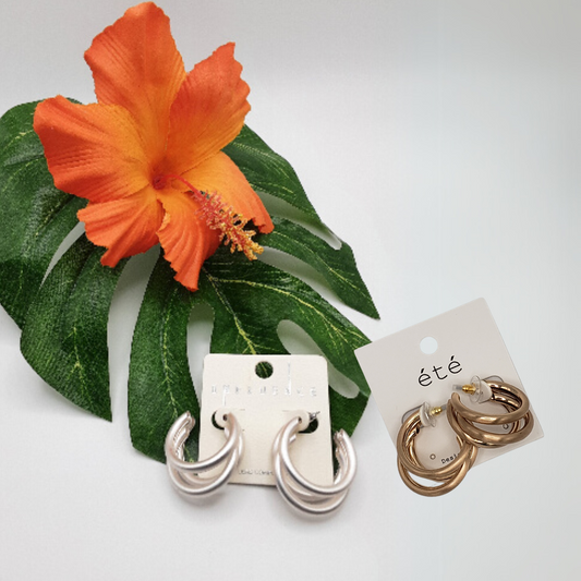 The SPECIAL EFFECTS Matte Metal Tube Hoop Earrings, with one matte silver pair resting on a leaf with an orange flower and a gleaming gold set beside a card, make a bold yet elegant statement perfect for jewelry lovers.