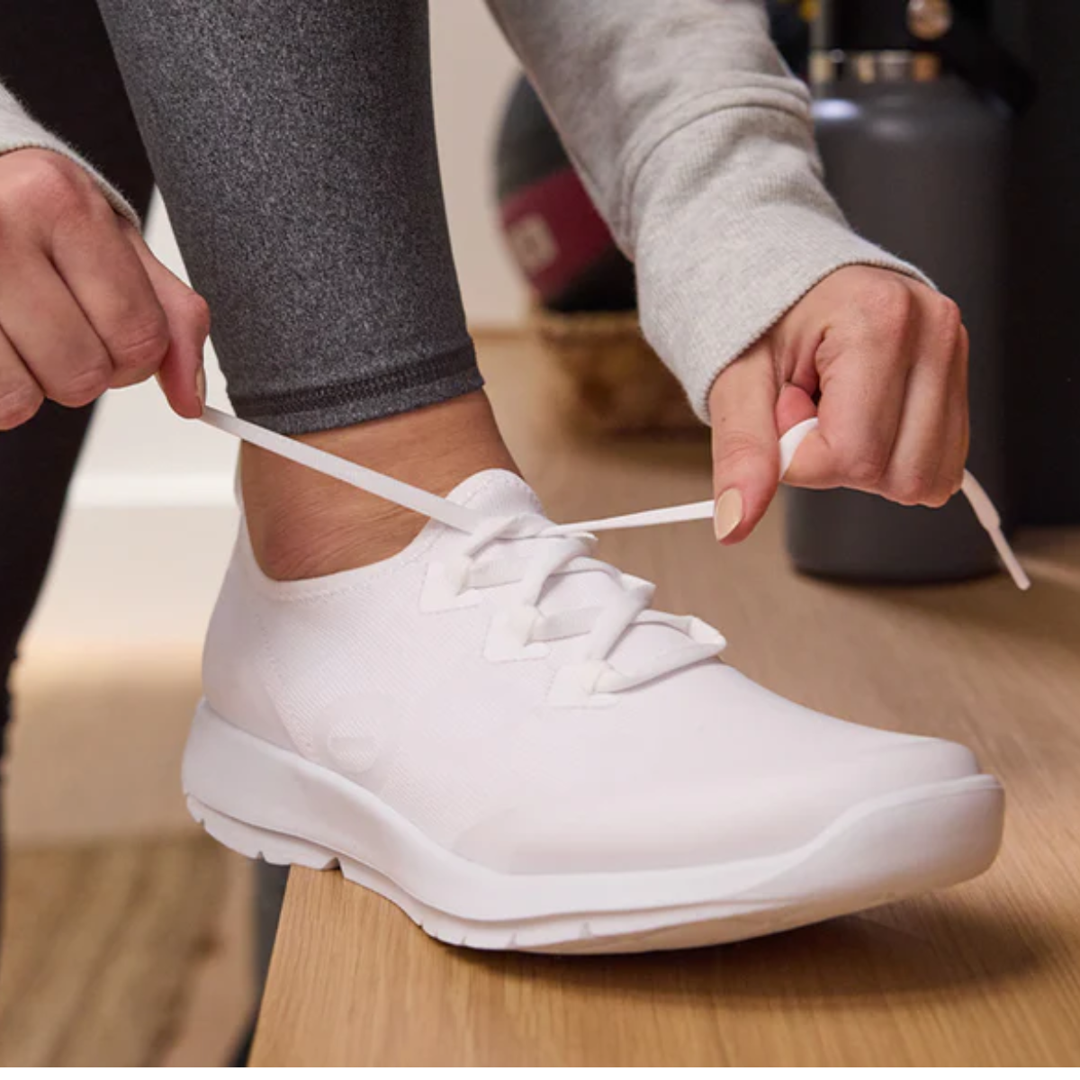 Someone in gray leggings ties their Flipflops & Whatnots Oofos Oomg Sport Lace shoes in White, featuring a FibreFlex upper, while sitting on a wooden surface—as ideal for active recovery.