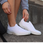 A person in white slip-on sneakers sits on concrete steps in a gray jacket, adjusting their Oomg Sport ladies OOfos in White—a perfect fit for an athlete's lifestyle.