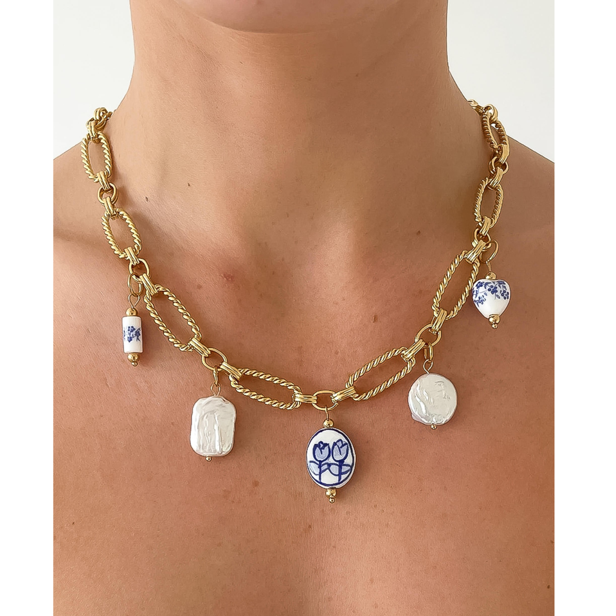 A person wearing a Flanerie 18K Gold Non-Tarnish Ceramic Floral Chain Necklace with exquisite blue and white hand-painted ceramic pendants of varying shapes and designs.