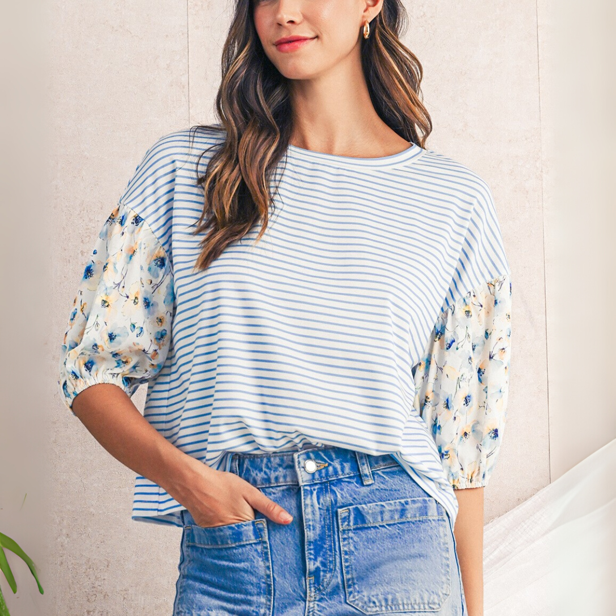 A woman indoors wears the Shewin Striped Floral Sleeve Pullover Top paired with high-waisted blue jeans, one hand in her pocket, showcasing a chic casual style.