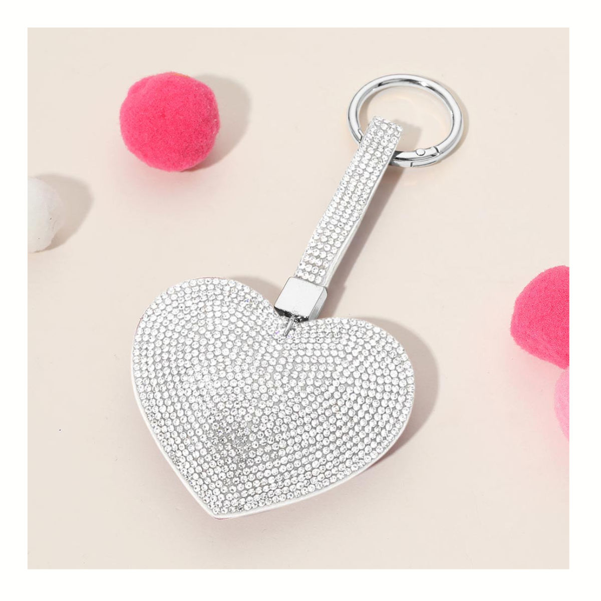 The Wona Trading Bling Heart Key Chain Bag Charm Assorted Colors features a silver heart with rhinestones, surrounded by three pink and one white pom-pom on a beige surface—making it a perfect sparkly accessory.