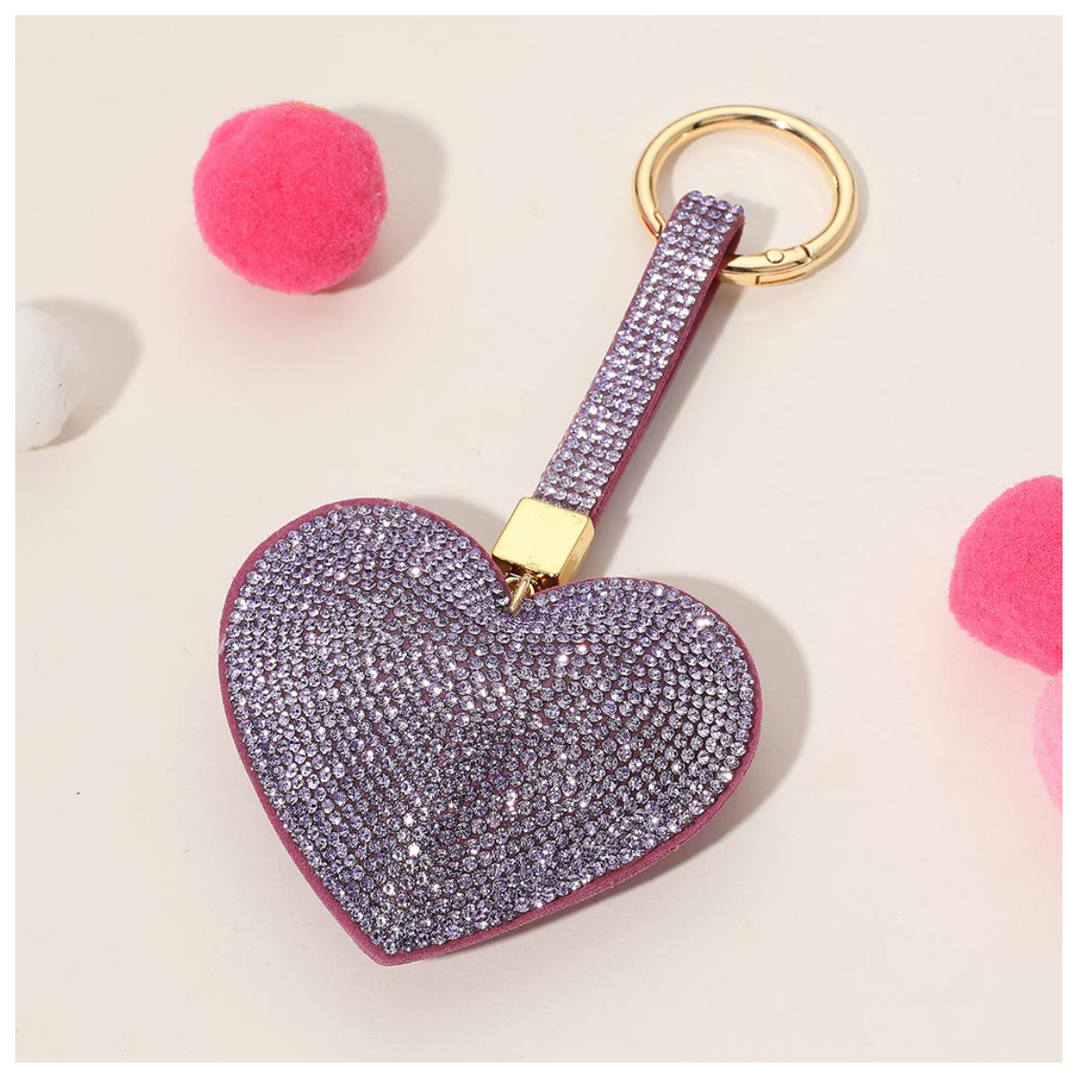 The Bling Heart Key Chain Bag Charm by Wona Trading features a sparkling heart with purple rhinestones, a gold keyring, and pink and white pom-poms, adding a fun accent to any accessory.