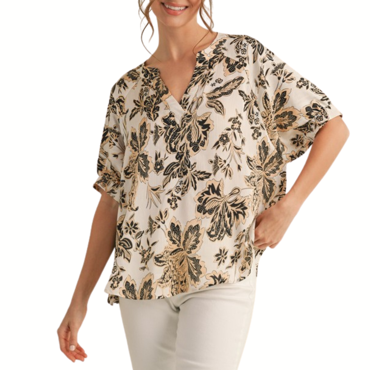 A woman wears the Fashiongo Flower Print Short Dolman Sleeves Top, a relaxed fit cream blouse with black and gold floral patterns, styled with light-colored pants.