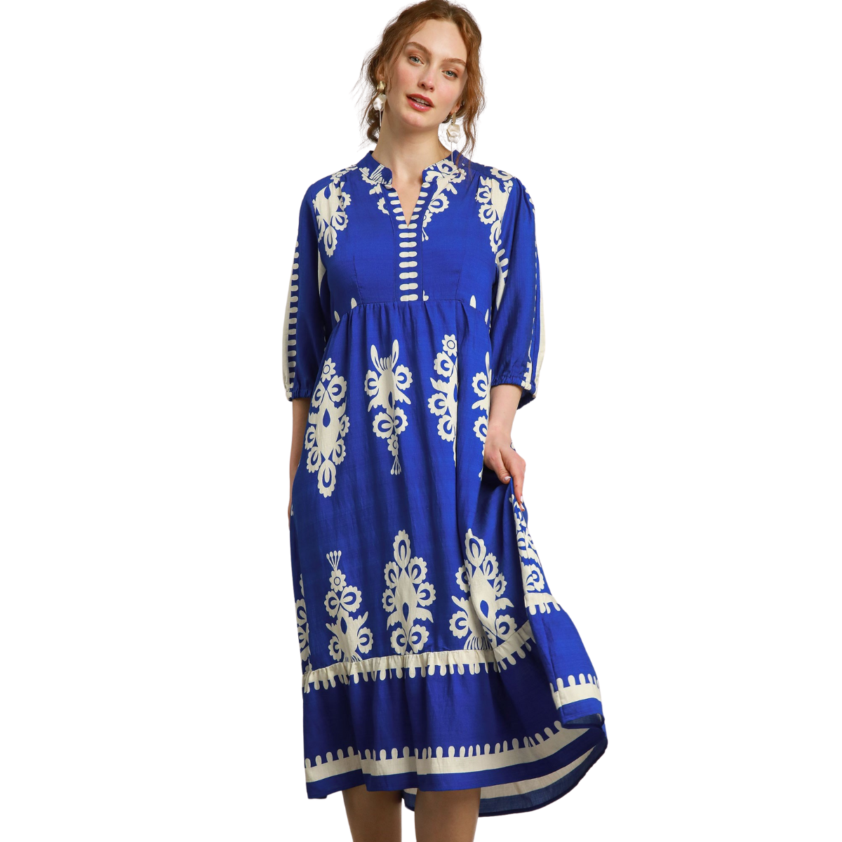 A woman wears the Umgee Desert Bloom Embroidered Midi Dress, featuring blue fabric with white floral patterns, a notched neckline, and three-quarter sleeves, standing with one hand in her pocket.
