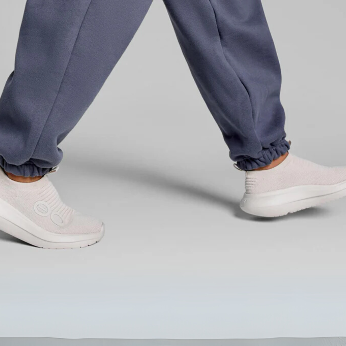 A person strolls in gray sweatpants and light gray slip-on shoes, exuding minimalist recovery. The OO My Zen in Chalk by OOFOS LLC features OOfoam™ technology, offering comfort and style against a plain backdrop.