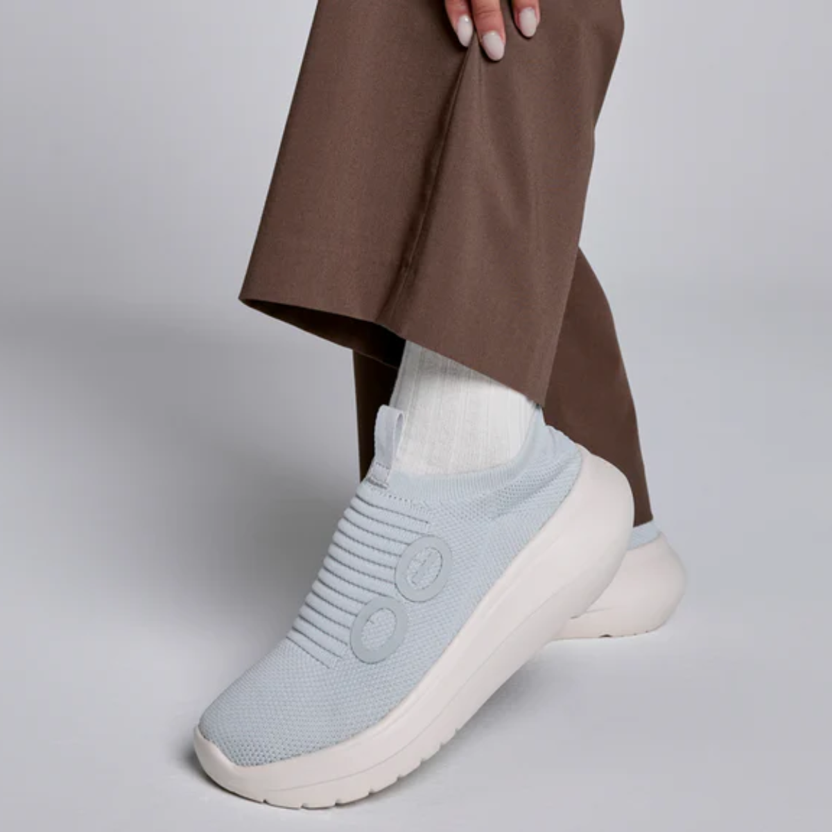 Wearing OOFOS LLC's OO My Zen in Sky, a light blue slip-on sneaker with a white sole and seamless circular knit design, someone pairs them with brown pants and white socks against a neutral background.