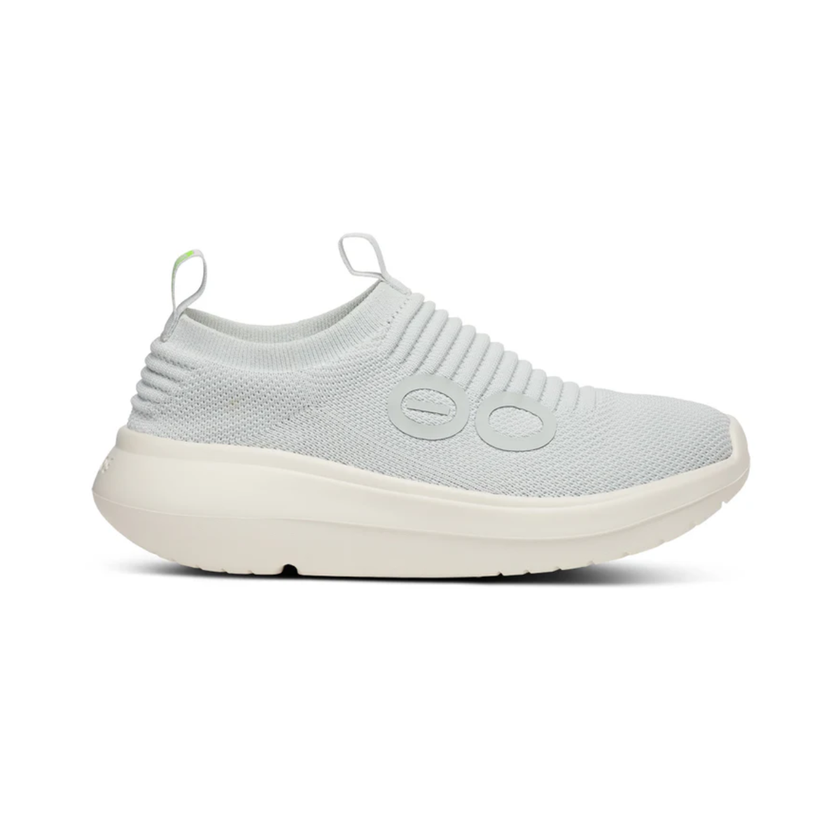 The OO My Zen in Sky by OOFOS LLC is a light grey slip-on sneaker with a rounded OOfoam sole, featuring a knitted upper with decorative ridges and circular side accents. Designed for active recovery, it includes pull tabs on the tongue and heel for easy wear.