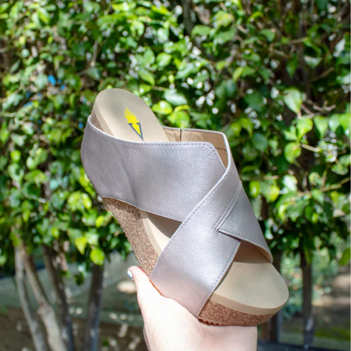 A Firefly Wedge in Pewter by Volatile, featuring crisscross straps and an ultra-comfort EVA insole for all-day comfort, is displayed against lush green foliage.